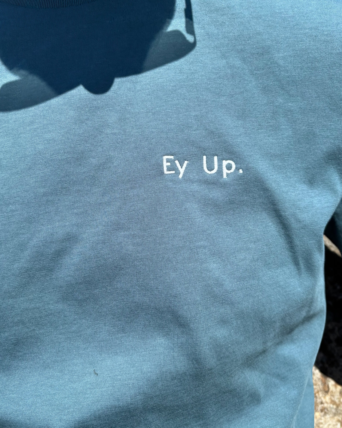 Close up of male model wearing Ey Up t-shirt top with embroidery slogan logo writing to left chest. 100% organic cotton t-shirt top, unisex clothing for men and women. Boxy fit, minimalist design. Yorkshire and Lancashire dialect.