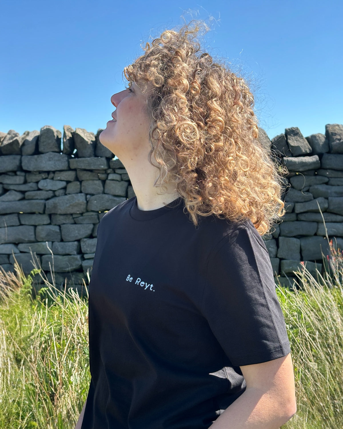 Female model wearing Be Reyt (be reight) t-shirt top in with slogan logo writing to left chest. 100% organic cotton t-shirt top, unisex clothing for men and women. Boxy fit, minimalist design. Yorkshire and Lancashire dialect.