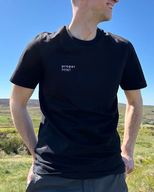 Male model wearing a black Proper Mint t-shirt top with white embroidery slogan logo writing to the right chest. 100% organic cotton t-shirt top, unisex clothing for men and women. Boxy fit, minimalist design. Yorkshire and Lancashire dialect.