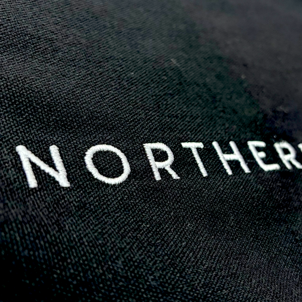 Close up of the stitching detail on a cotton canvas tote shoulder bag - perfect eco conscious shopping bag / bag for life. Great gift for Northerners and people from Yorkshire of Lancashire. bag with embroidered Northerner slogan in bold, minimalist font. Bag shown laying flat on the ground with a background. Men's and ladies / women's bag