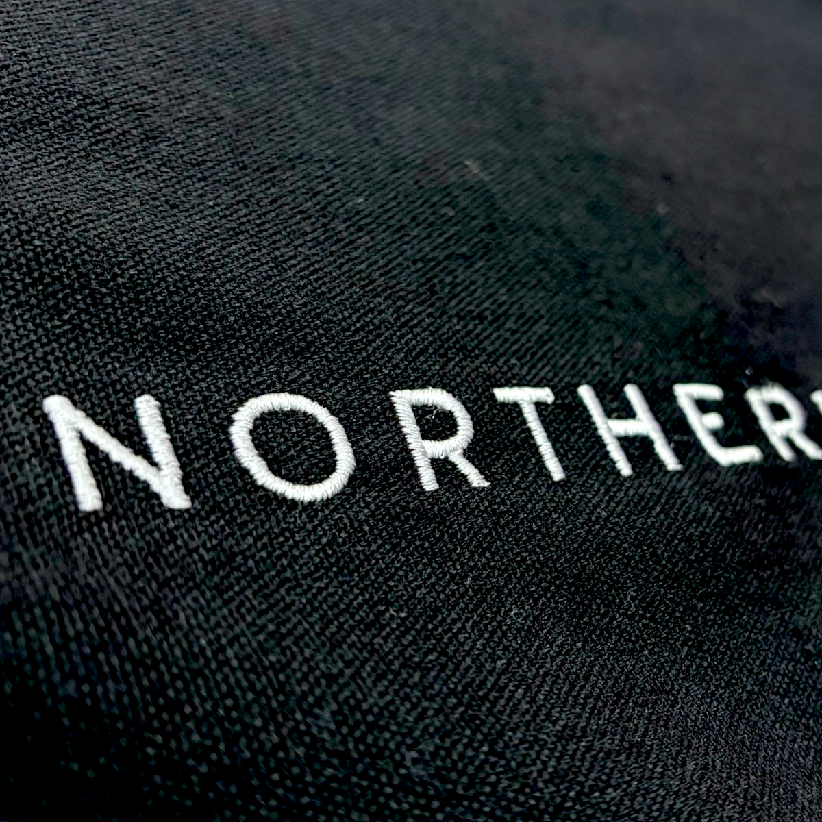 Close up of the stitching detail on a cotton canvas tote shoulder bag - perfect eco conscious shopping bag / bag for life. Great gift for Northerners and people from Yorkshire of Lancashire. bag with embroidered Northerner slogan in bold, minimalist font. Bag shown laying flat on the ground with a background. Men's and ladies / women's bag