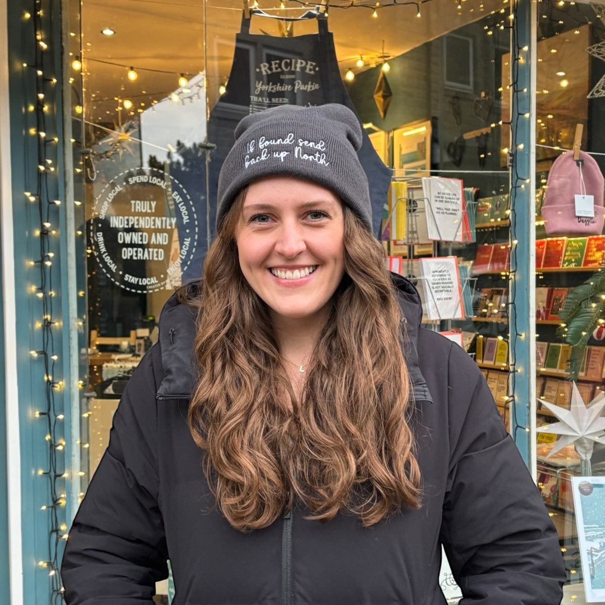 If found send back up North Northerner Yorkshire Lancashire funny Yorkshire Northern humour dialect gift - beanie hat to stay warm in cold weather and Winter. embroidered handwriting font logo with star to the front cuff of the beanie. Dark hat. Female model with long wavy mousy hair looking at the camera. Christmas gift. British humour gift. Unisex hat for women ladies and men. Model is standing in front of a shop window.