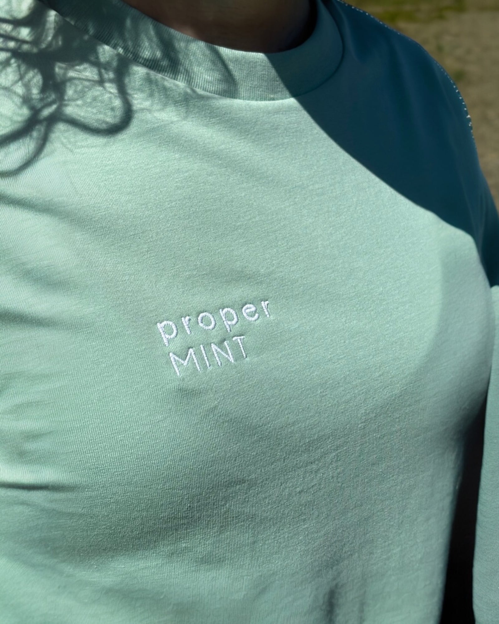 Female model wearing a light cream white neutral Proper Mint t-shirt top with black embroidery slogan logo writing to the right chest. 100% organic cotton t-shirt top, unisex clothing for men and women. Boxy fit, minimalist design. Yorkshire and Lancashire dialect.