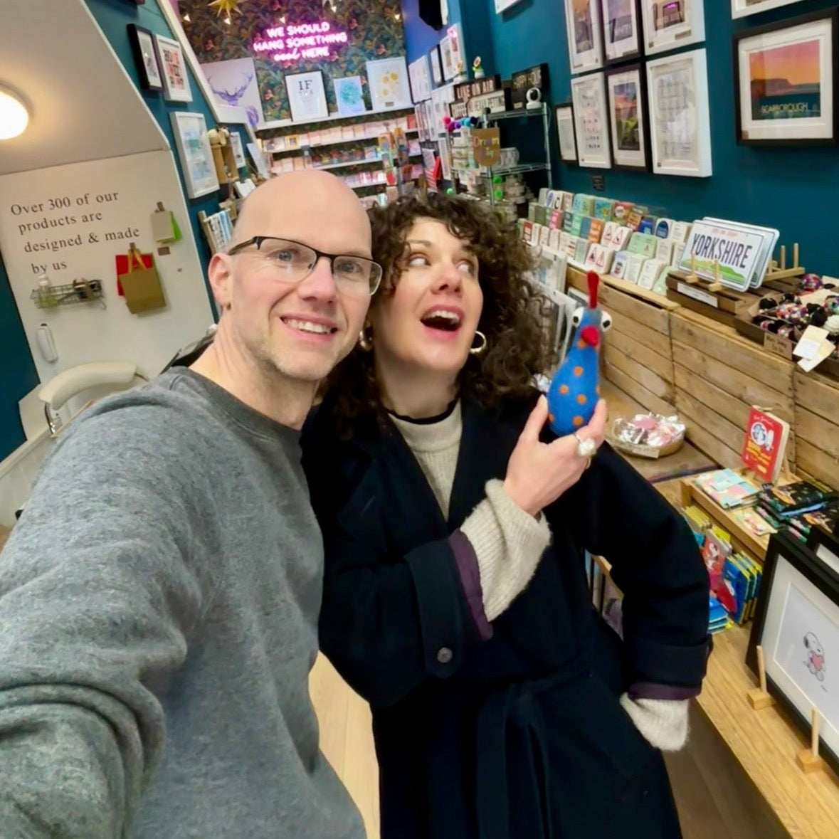 A man and a woman taking a selfie inside a retail shop. The woman is wearing a dark navy blue coat over a beige top and holding a small blue and red chicken