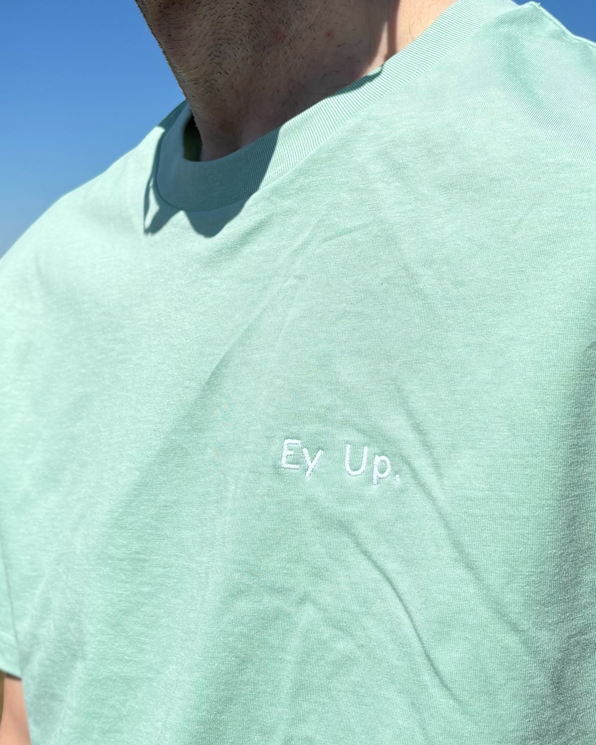 Close up of male model wearing minty fresh light Ey Up t-shirt top with embroidery slogan logo writing to left chest. 100% organic cotton t-shirt top, unisex clothing for men and women. Boxy fit, minimalist design. Yorkshire and Lancashire dialect.