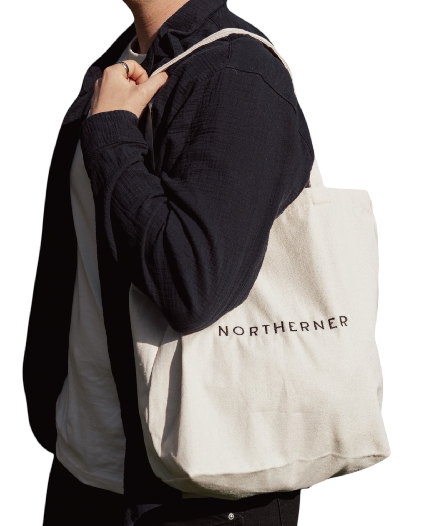 Cotton canvas cream natural tote shoulder bag - perfect eco conscious shopping bag / bag for life. Great gift for Northerners and people from Yorkshire of Lancashire. Cream bag with black embroidered Northerner slogan in bold, minimalist font. Bag shown worn on the shoulder of a male model wearing a shirt over a t-shirt. Men's and ladies / women's bag