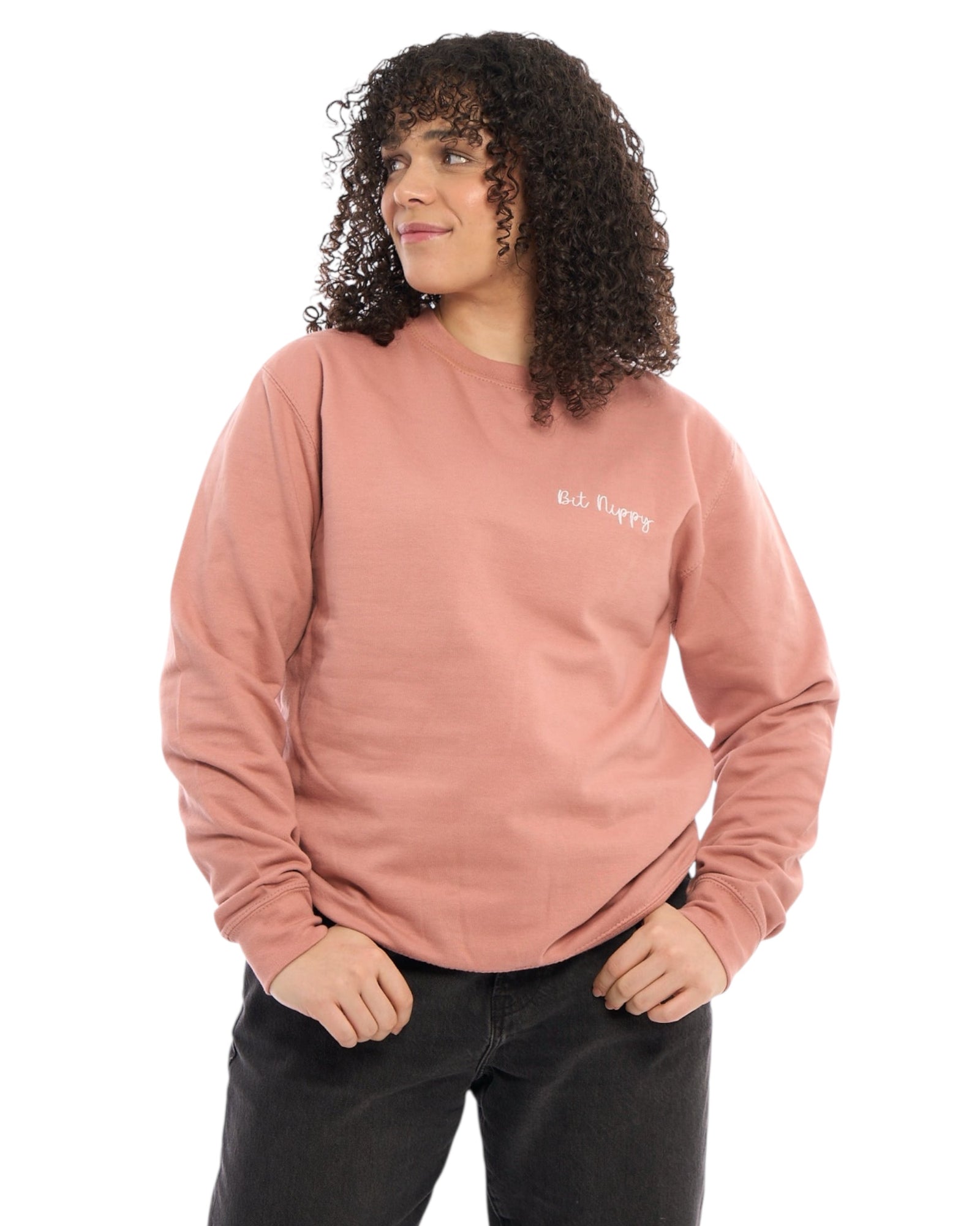 Premium quality designer embroidered embroidery Bit Nippy cold weather slogan logo crew neck long sleeve sweatshirt. Light sage green top with subtle white minimalist small logo on the left breast. Yorkshire Lancashire dialect. Funny local slogan logo dialect. Men's and women's clothing - unisex wear. Female model smiling with curly shoulder length hair.