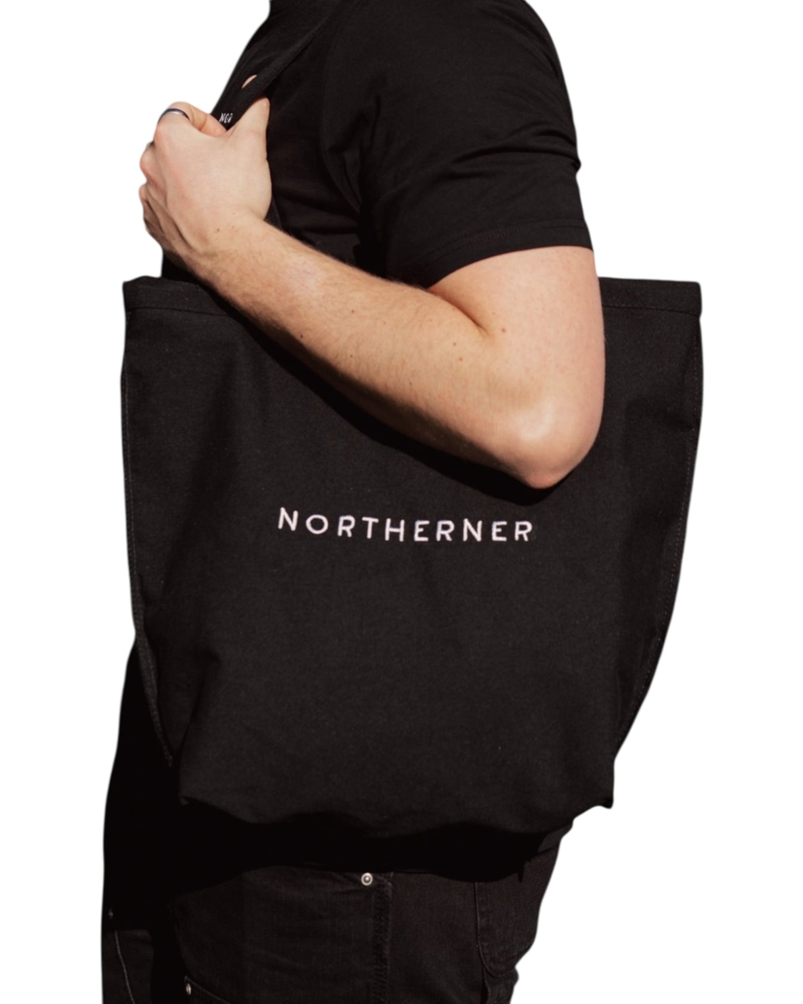 Cotton canvas cream natural tote shoulder bag - perfect eco conscious shopping bag / bag for life. Great gift for Northerners and people from Yorkshire of Lancashire. Cream bag with black embroidered Northerner slogan in bold, minimalist font. Bag shown worn on the shoulder of a male model wearing a shirt over a t-shirt. Men's and ladies / women's bag