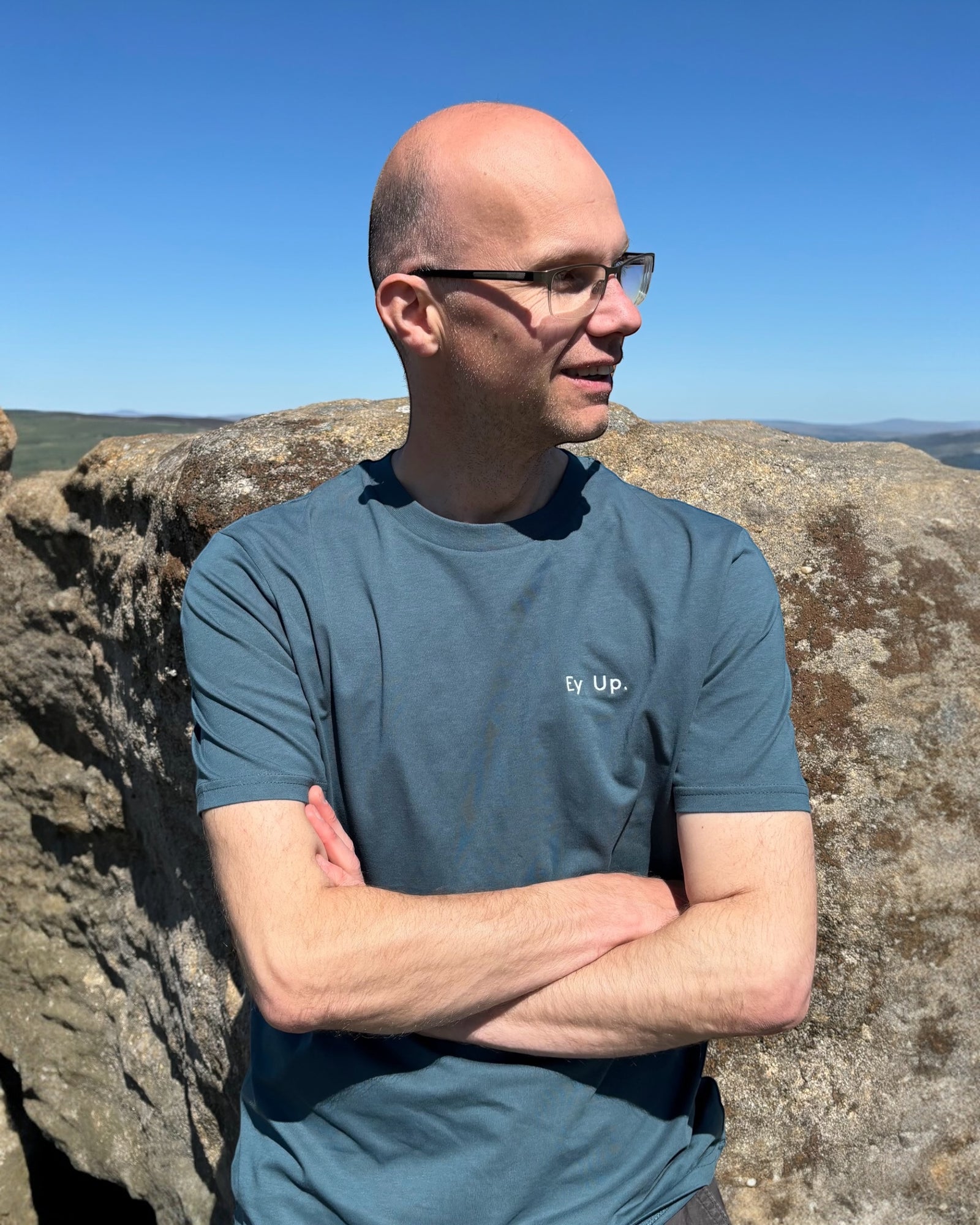 Male model wearing teal blue green Ey Up t-shirt top with white embroidery slogan logo writing to left chest. 100% organic cotton t-shirt top, unisex clothing for men and women. Boxy fit, minimalist design. Yorkshire and Lancashire dialect. 