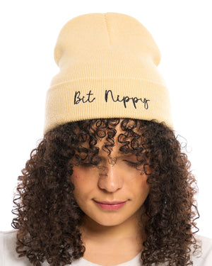Bit Nippy funny Yorkshire Northerner humour dialect gift - beanie hat to stay warm in cold weather and Winter. Black embroidered handwriting font logo to the front cuff of the beanie. Beige stone neutral cream hat. Female model looking down with curly ringlet hair. British humour gift. Unisex hat for men and women.