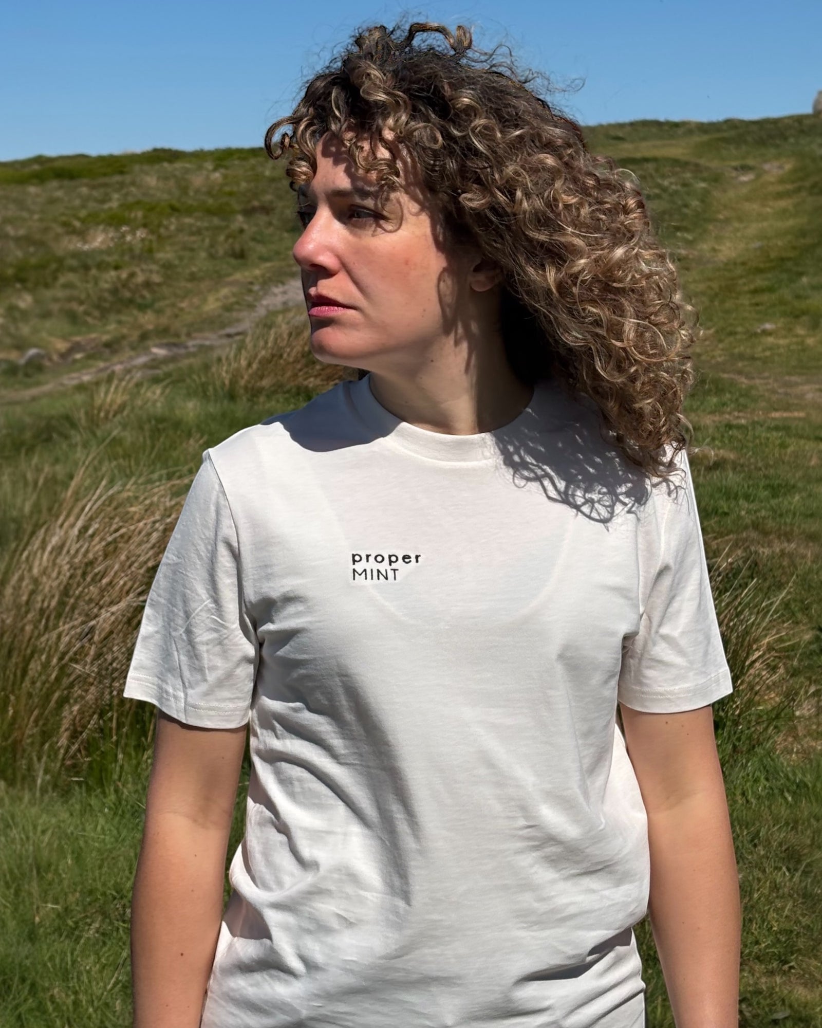 Female model wearing a light cream white neutral Proper Mint t-shirt top with black embroidery slogan logo writing to the right chest. 100% organic cotton t-shirt top, unisex clothing for men and women. Boxy fit, minimalist design. Yorkshire and Lancashire dialect.