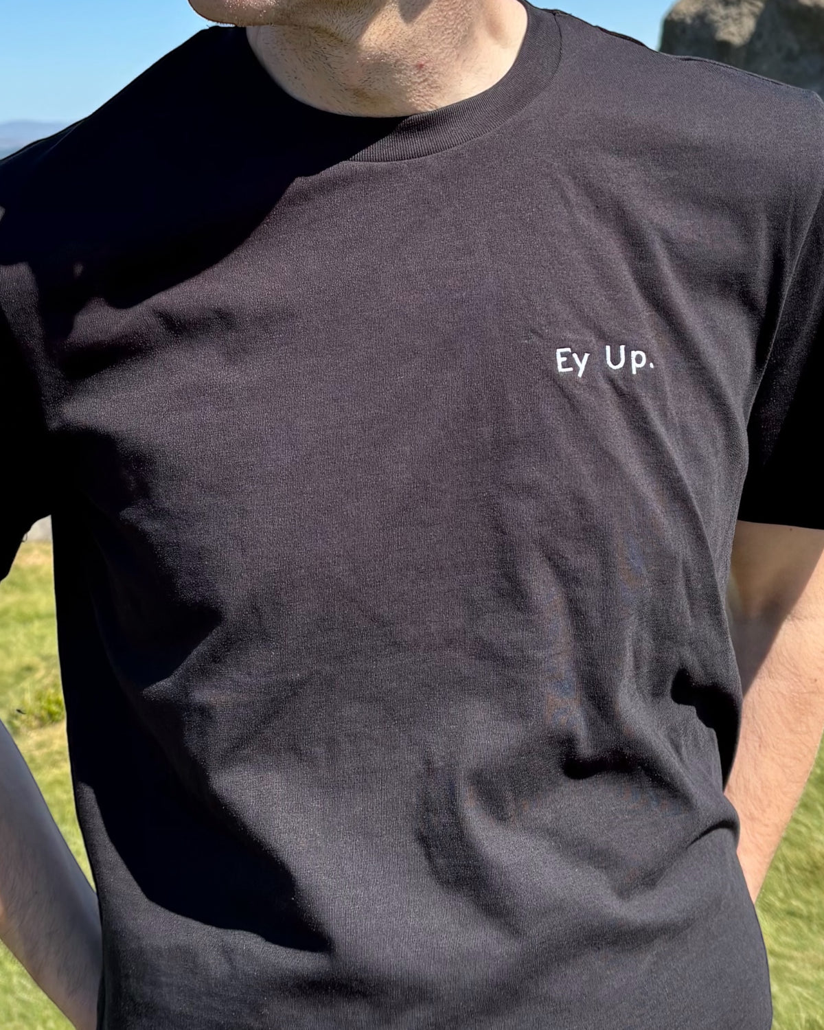 Close up of male model wearing Ey Up t-shirt top with embroidery slogan logo writing to left chest. 100% organic cotton t-shirt top, unisex clothing for men and women. Boxy fit, minimalist design. Yorkshire and Lancashire dialect.