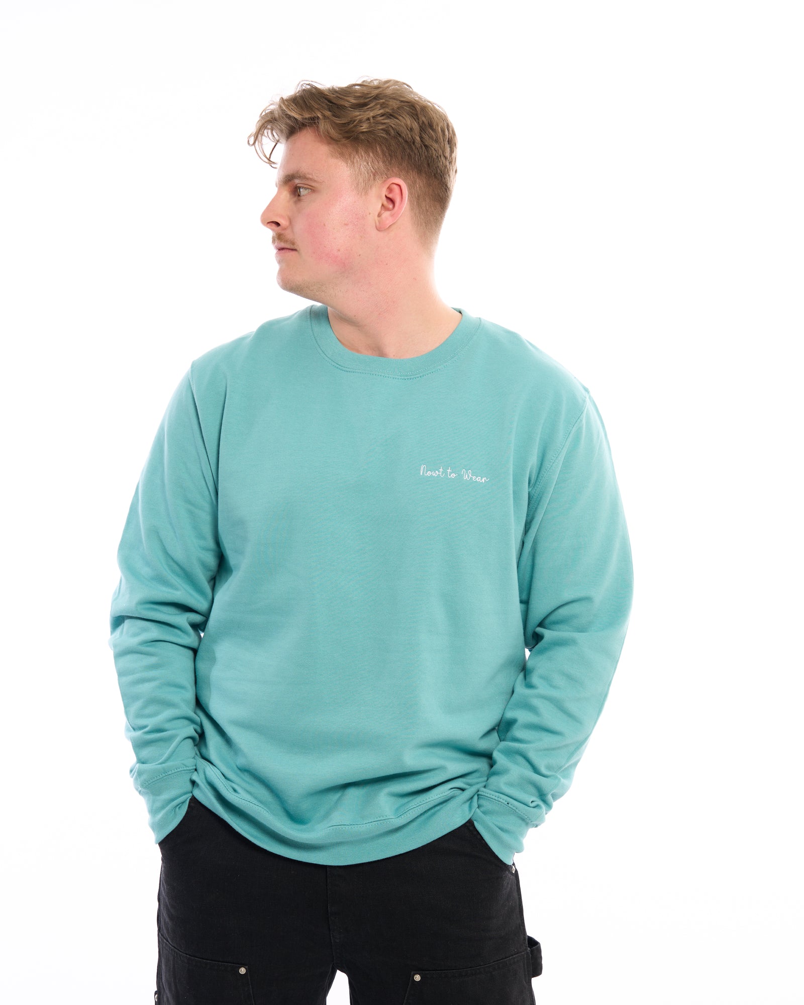 Premium quality designer embroidered embroidery Nowt to Wear (nothing to wear) funny slogan logo crew neck long sleeve sweatshirt. Turquoise teal blue green top with subtle handwriting white minimalist small logo on the left breast. Yorkshire Lancashire dialect. Funny local slogan logo dialect. Men's and women's clothing - unisex wear. Male model with hand in pocket looking to the right.