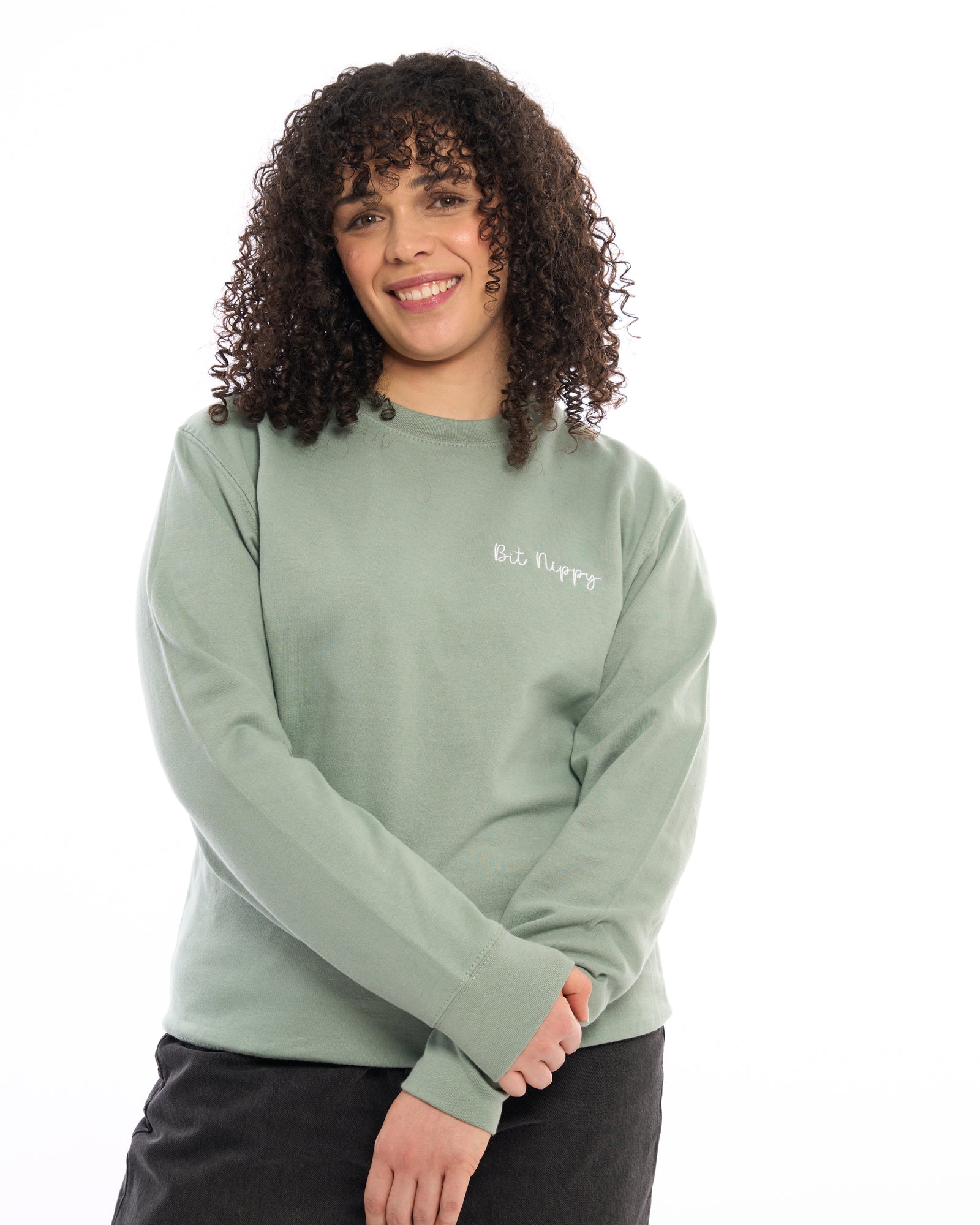 Premium quality designer embroidered embroidery Bit Nippy cold weather slogan logo crew neck long sleeve sweatshirt. Light sage green top with subtle white minimalist small logo on the left breast. Yorkshire Lancashire dialect. Funny local slogan logo dialect. Men's and women's clothing - unisex wear. Female model smiling with curly shoulder length hair.