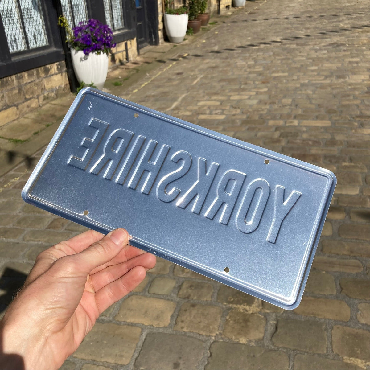 LighthouseLane embossed aluminium Yorkshire USA style licence plate held in hand against a cobbled street background