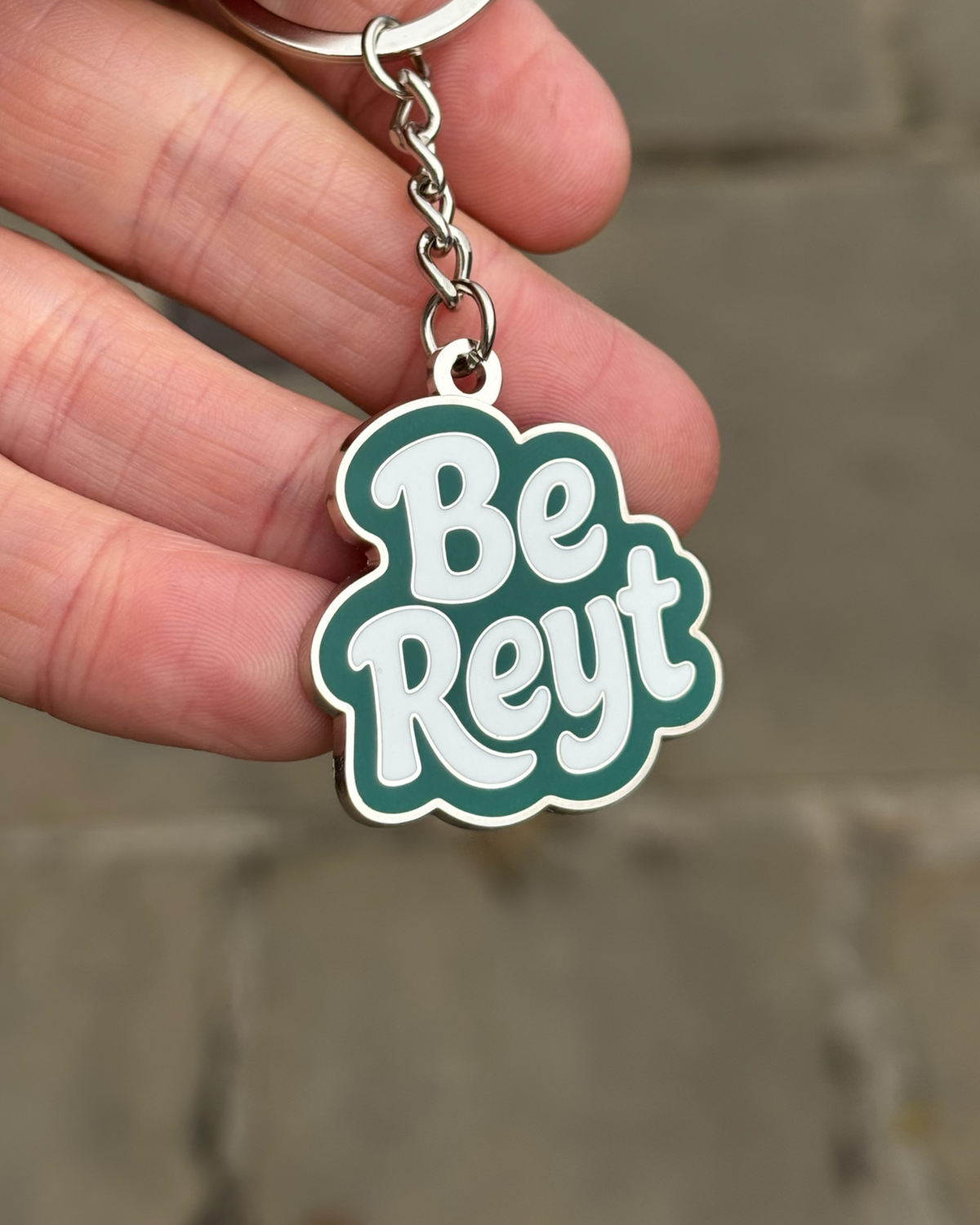 LighthouseLane Be Reyt enamel keyring in deep green with white text held in a hand