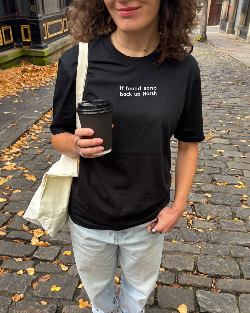 LighthouseLane black organic cotton unisex t-shirt with If Found Send Up North embroidery worn on a cobblestone street