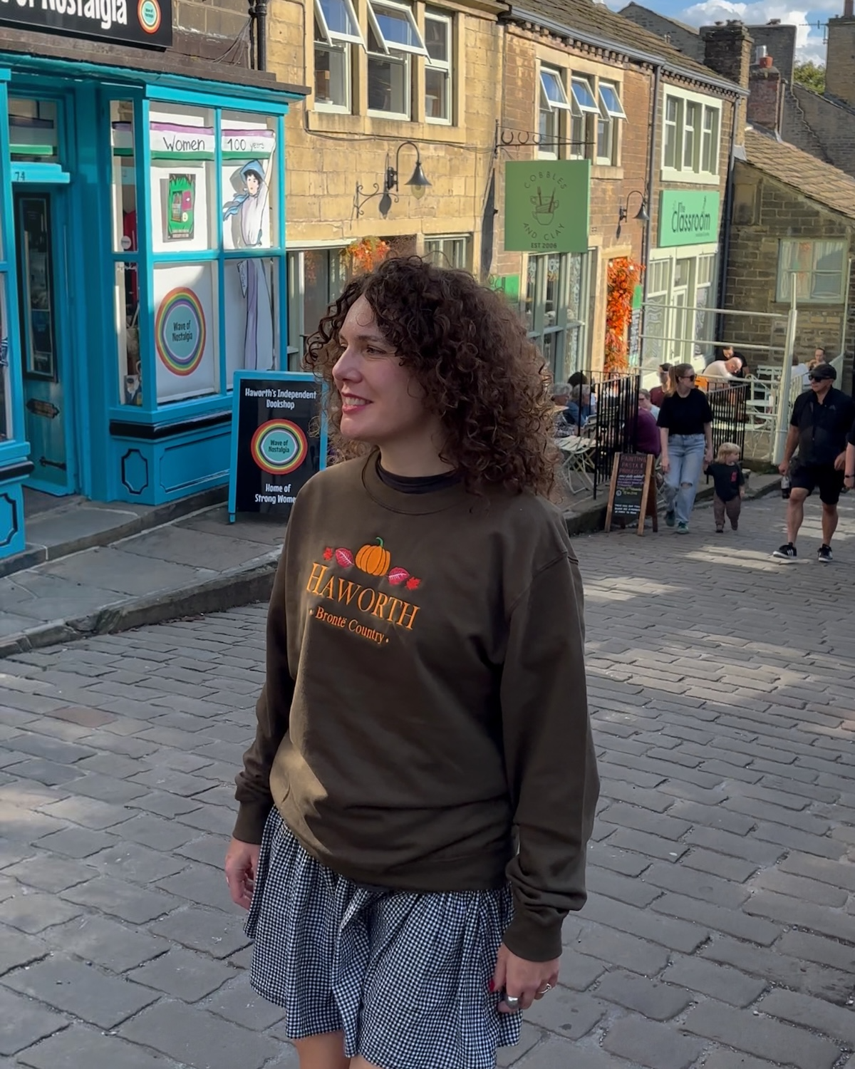 LighthouseLane unisex olive green sweatshirt with embroidered Haworth pumpkin and autumn leaf design on a cobbled street