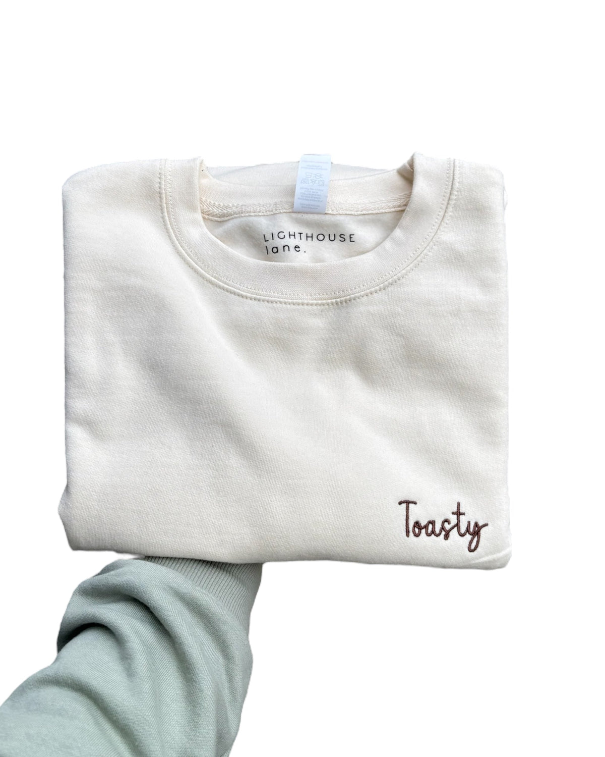 LighthouseLane cream cotton blend sweatshirt with Toasty embroidered slogan, folded and held by a hand.